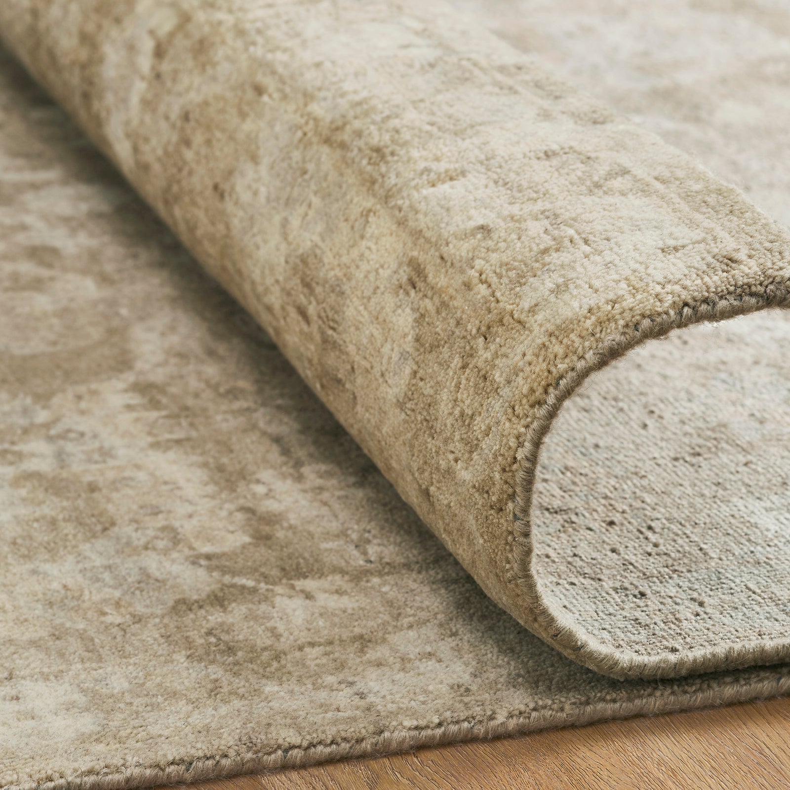 Close-up of the Remus hand-knotted wool rug rolled at the edge, showing the dense wool pile construction, cotton foundation backing, and natural textural variation in the ivory and taupe fibers