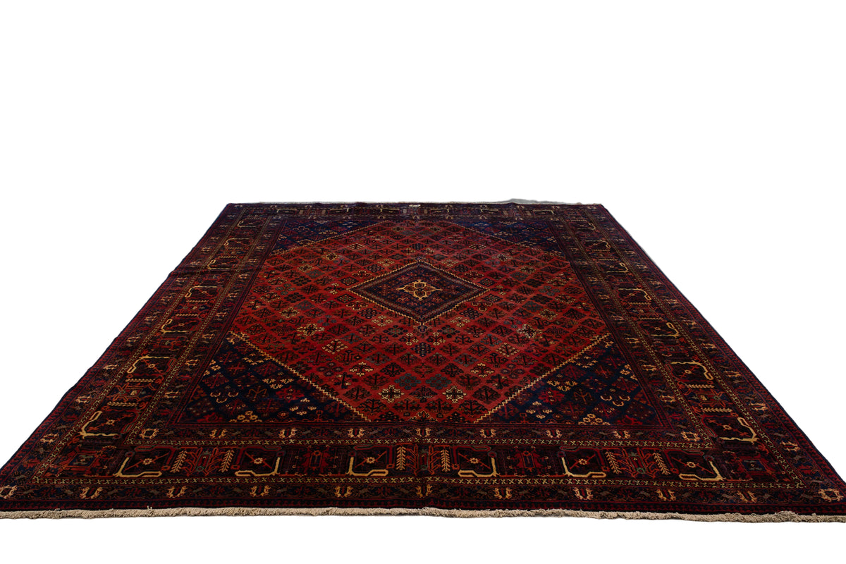 A large traditional Persian-style area rug with a dark red central field, navy blue corners, and beige geometric borders, displayed on a light wood floor inside a carpet showroom.